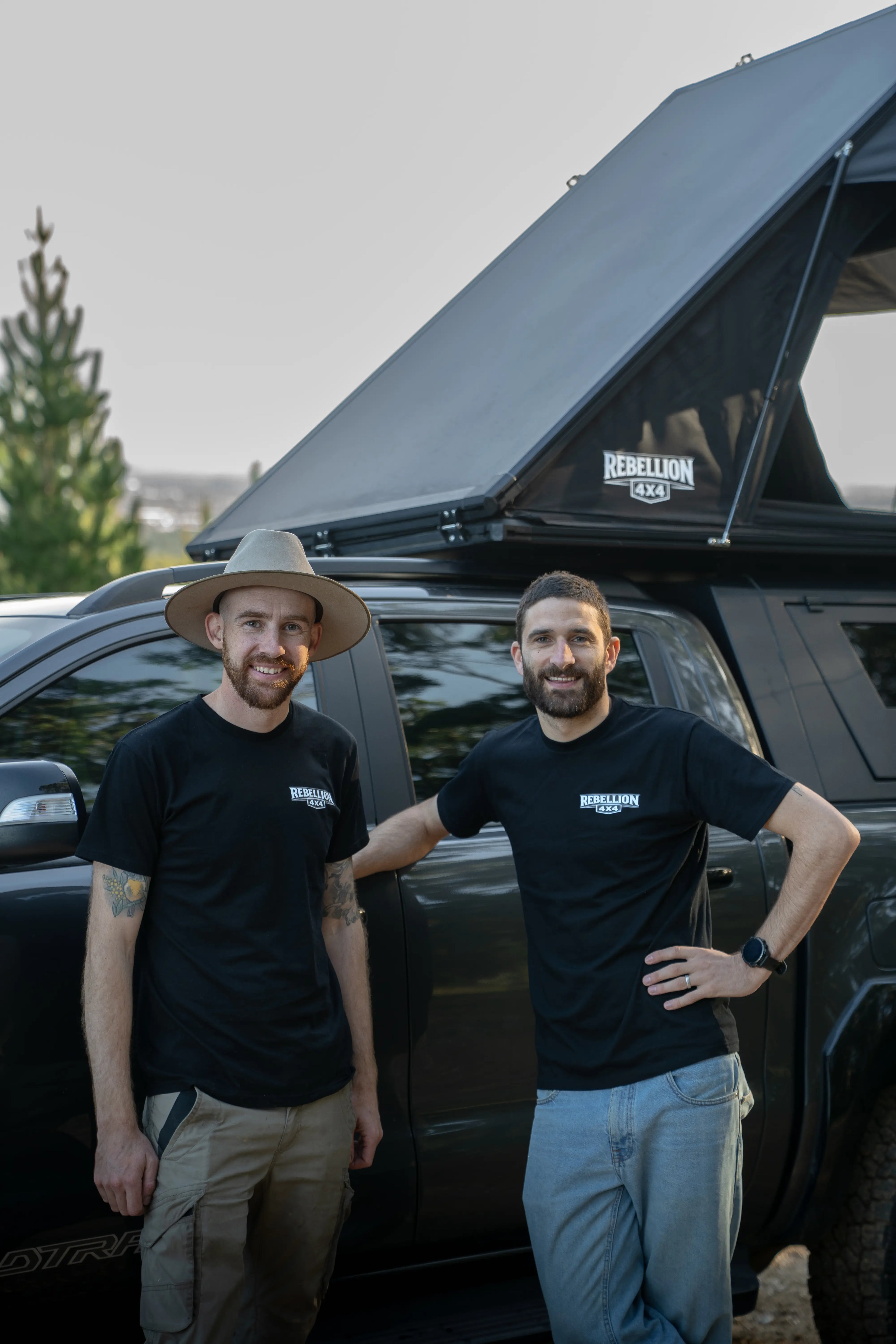 Two men standing next to a black truck with a rooftop tent, smiling at the camera.
