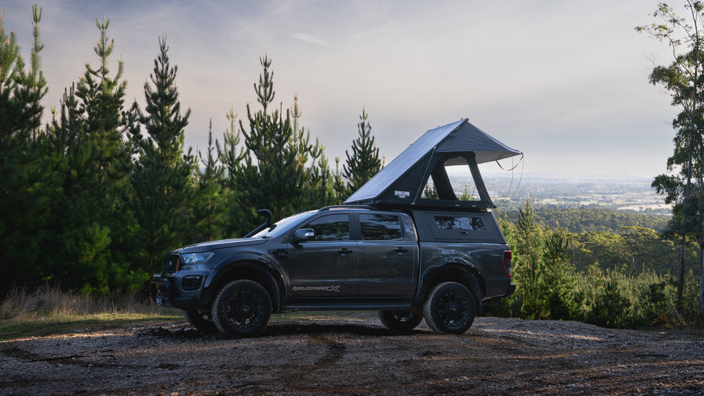 Dark gray truck with a rooftop tent parked on a dirt road surrounded by trees.