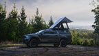 Dark gray truck with a rooftop tent parked on a dirt road surrounded by trees.