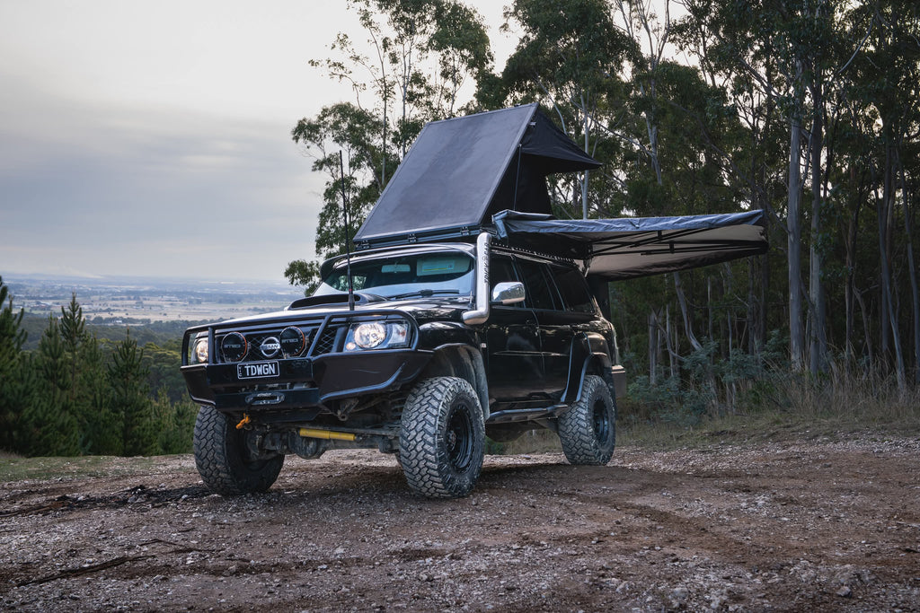 Black off-road vehicle with a roof tent parked on a dirt road surrounded by trees.