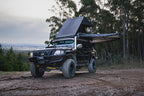 Black off-road vehicle with a roof tent parked on a dirt road surrounded by trees.