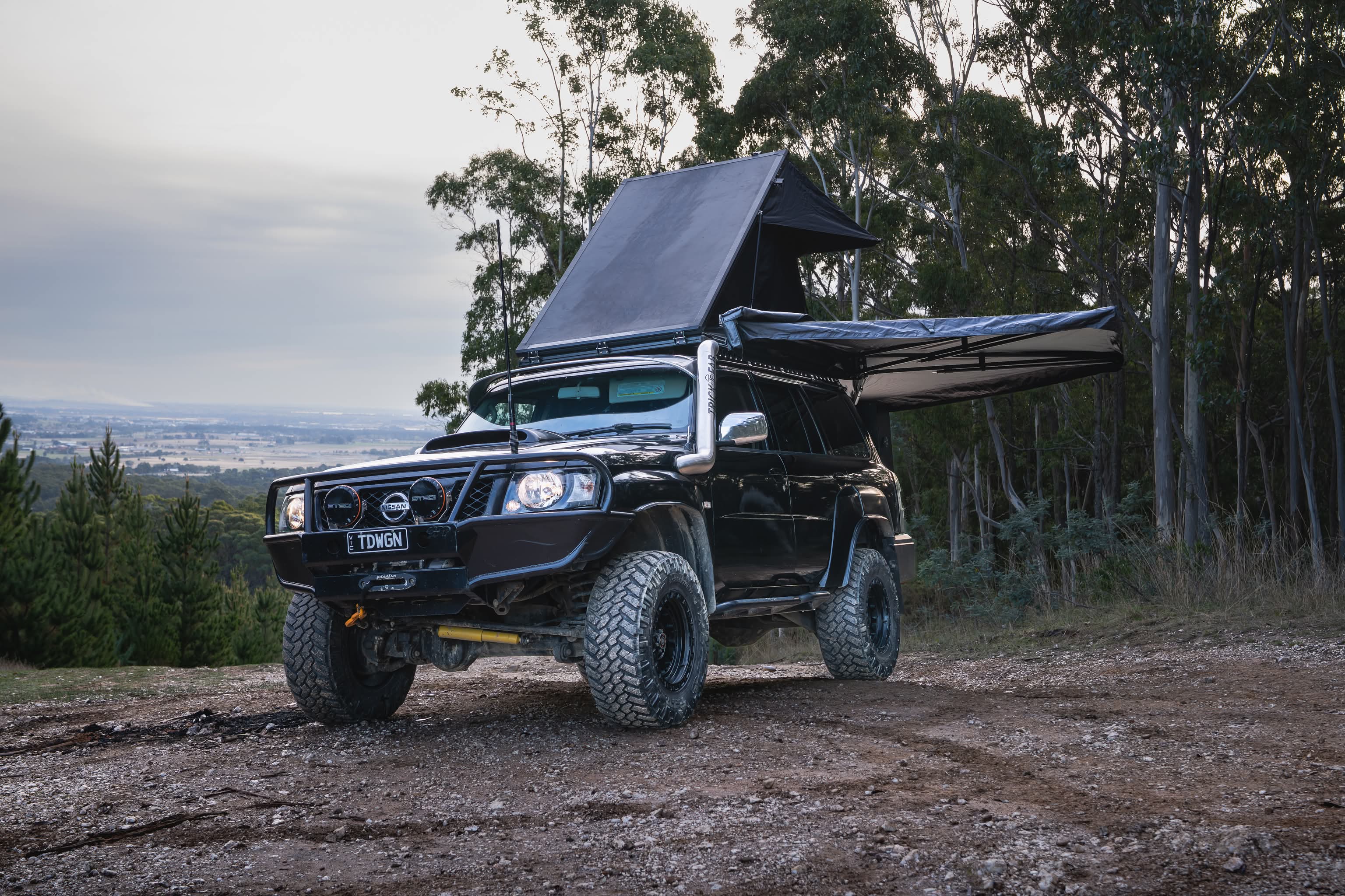 Black off-road vehicle with a roof tent parked on a dirt road surrounded by trees.