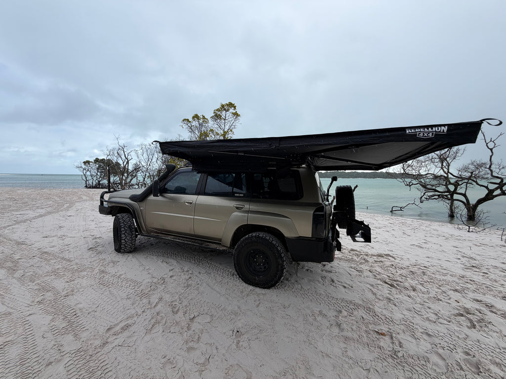 A beige off-road SUV parked on a sandy beach with a large black awning extended over the roof, under an overcast sky.