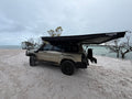 A beige off-road SUV parked on a sandy beach with a large black awning extended over the roof, under an overcast sky.