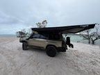 A beige off-road SUV parked on a sandy beach with a large black awning extended over the roof, under an overcast sky.