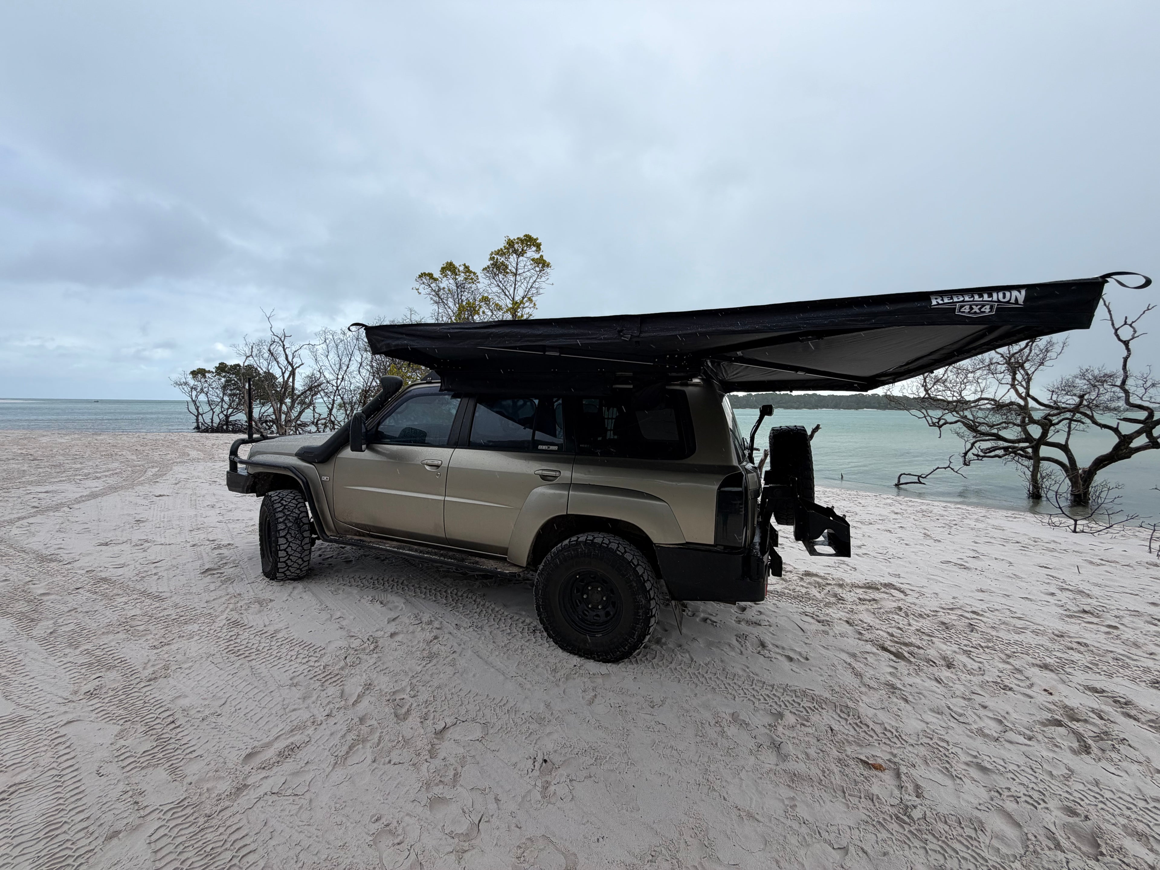 A beige off-road SUV parked on a sandy beach with a large black awning extended over the roof, under an overcast sky.