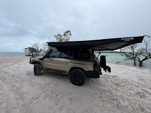 A beige off-road SUV parked on a sandy beach with a large black awning extended over the roof, under an overcast sky.