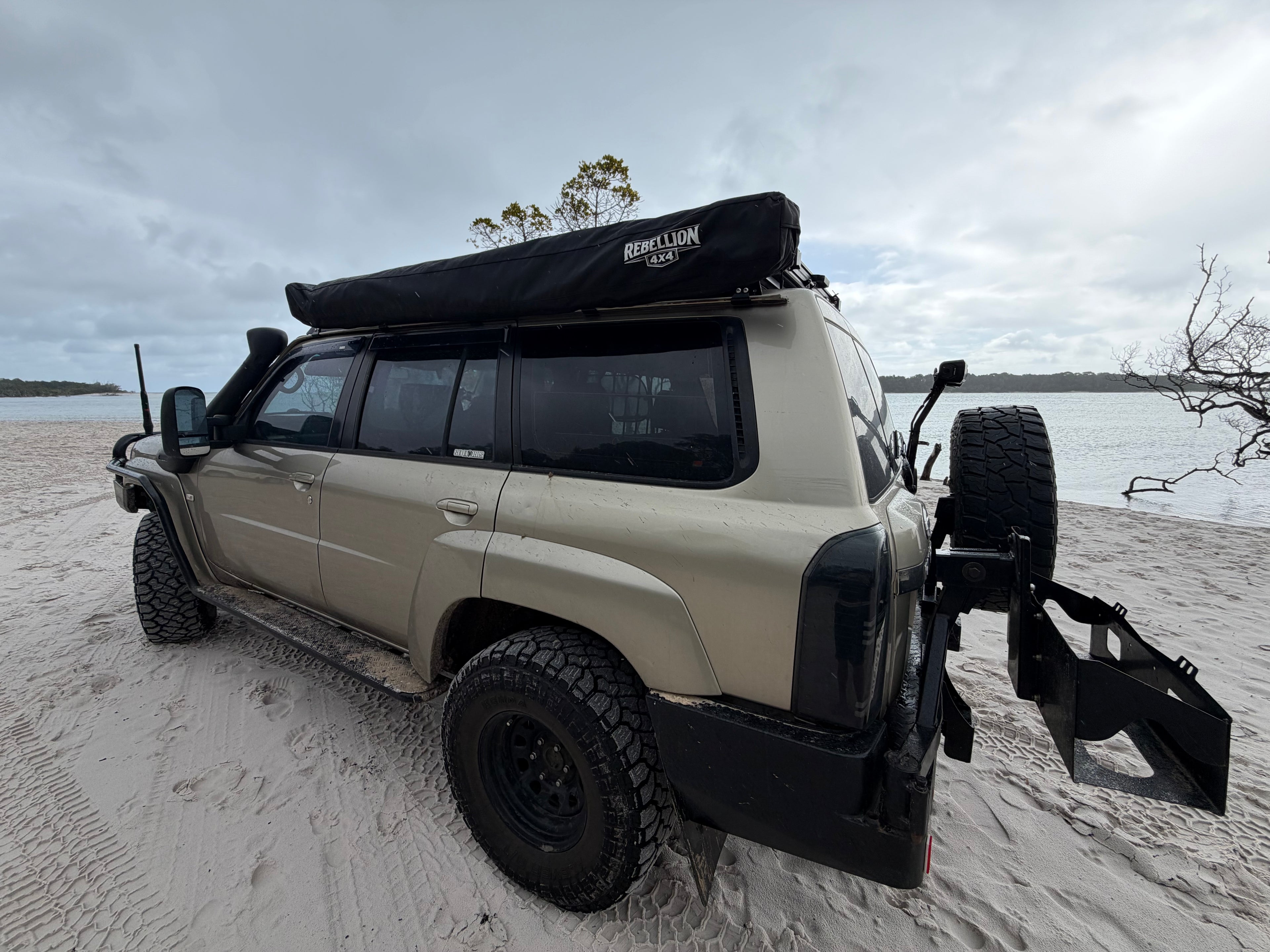 Beige Nissan patrol with black awning parked on sandy beach with overcast sky