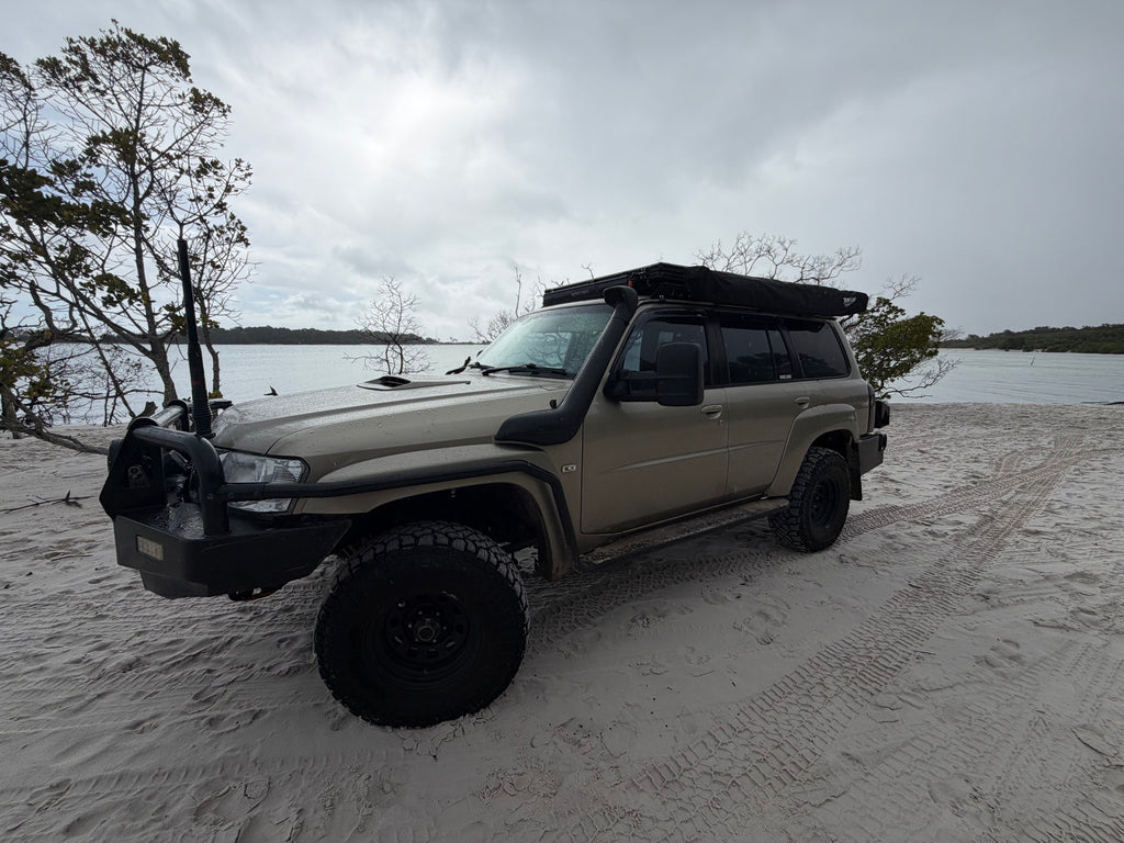 Beige Nissan Patrol parked on Sandy beach with cloudy skies with black awning and trees in the backround