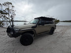 Beige Nissan Patrol parked on Sandy beach with cloudy skies with black awning and trees in the backround