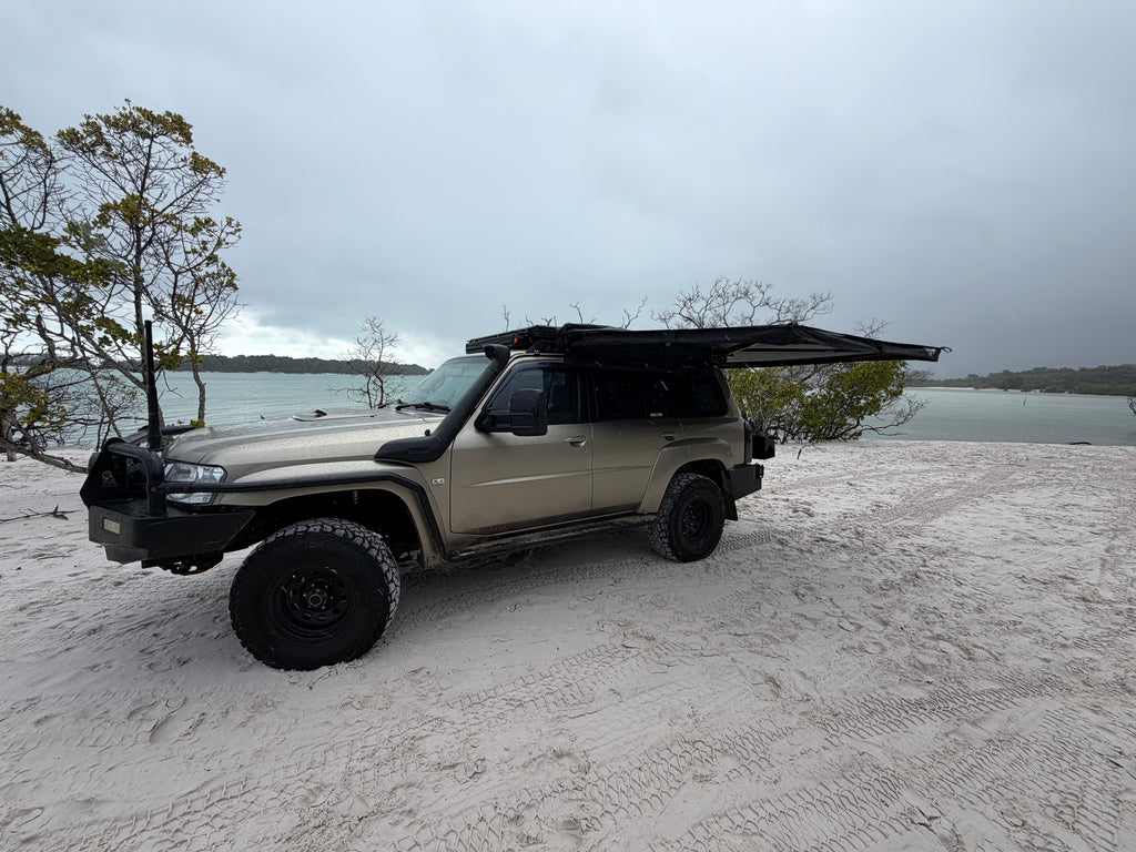 A beige off-road SUV parked on a sandy beach with a black pop-up canopy tent mounted over the roof, overlooking a calm body of water under an overcast sky.