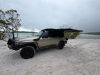 A beige off-road SUV parked on a sandy beach with a black pop-up canopy tent mounted over the roof, overlooking a calm body of water under an overcast sky.