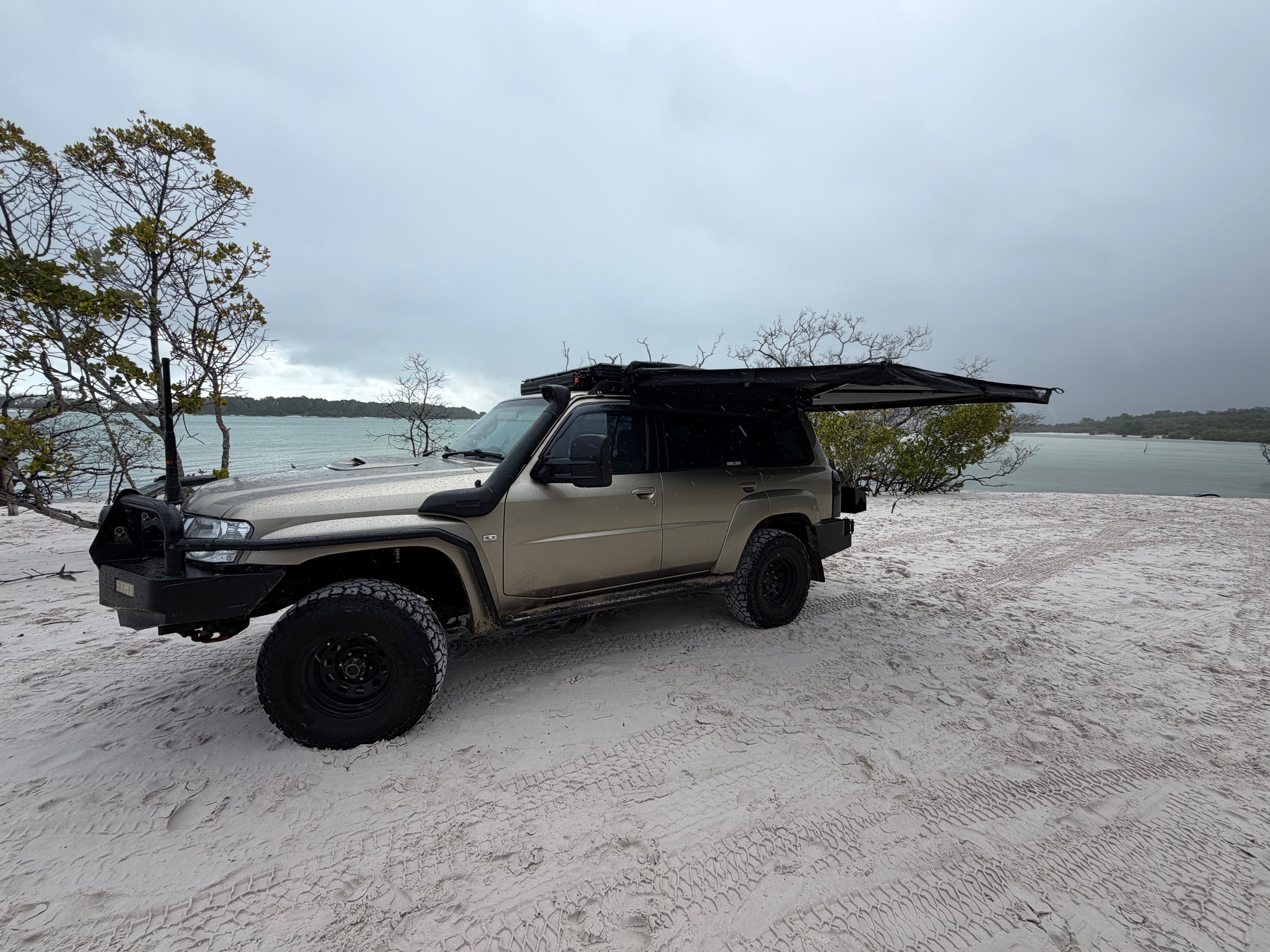 A beige off-road SUV parked on a sandy beach with a black pop-up canopy tent mounted over the roof, overlooking a calm body of water under an overcast sky.