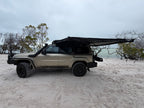 A beige off-road SUV parked on white sand under an overcast sky, with a black awning extended over the roof and trees in the background.