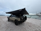 A beige off-road SUV parked on a sandy beach with a large black rooftop canopy tent extended over the vehicle, with water and cloudy sky in the background.