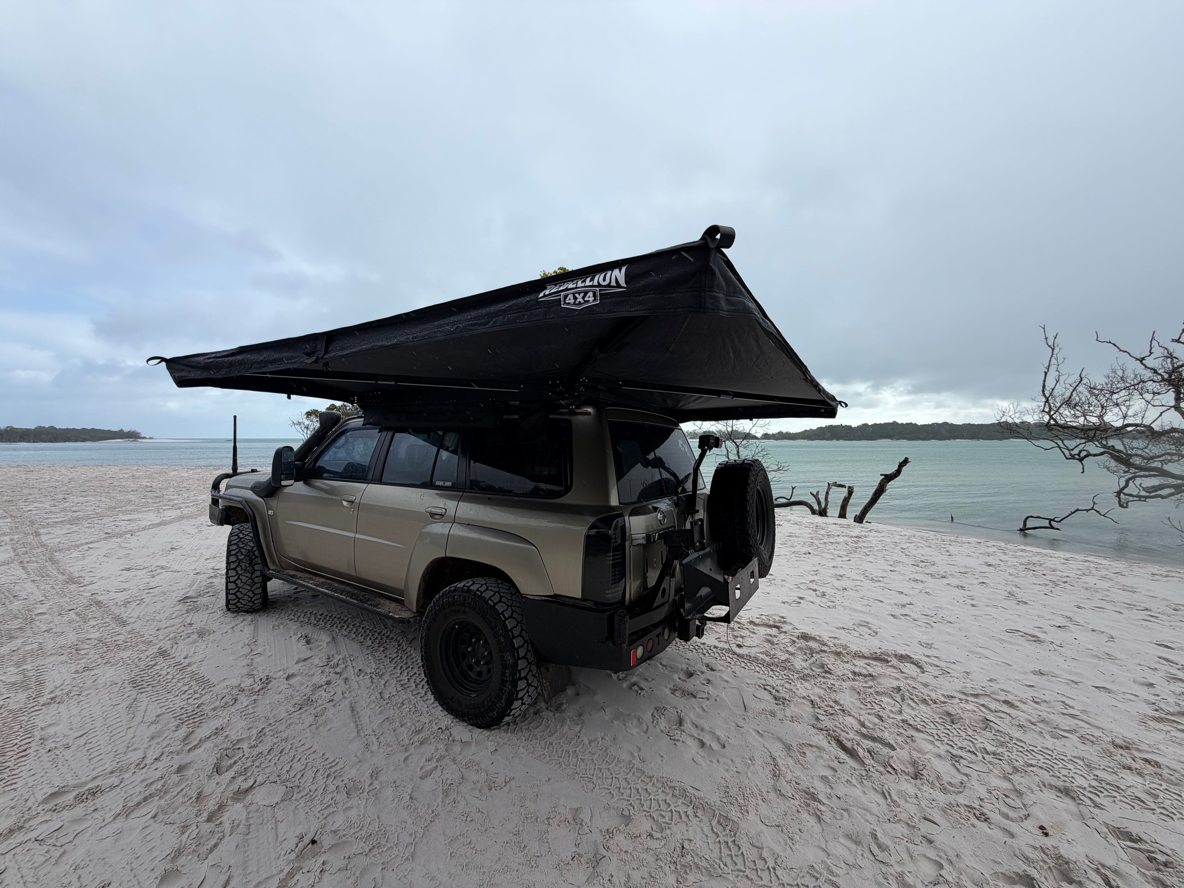A beige off-road SUV parked on a sandy beach with a large black rooftop canopy tent extended over the vehicle, with water and cloudy sky in the background.