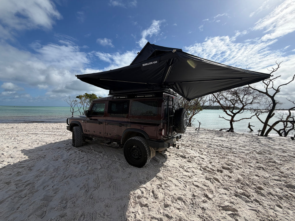 A dark red off-road vehicle parked on a sandy beach with a large black rooftop tent canopy extended over the vehicle, under a blue sky with clouds.