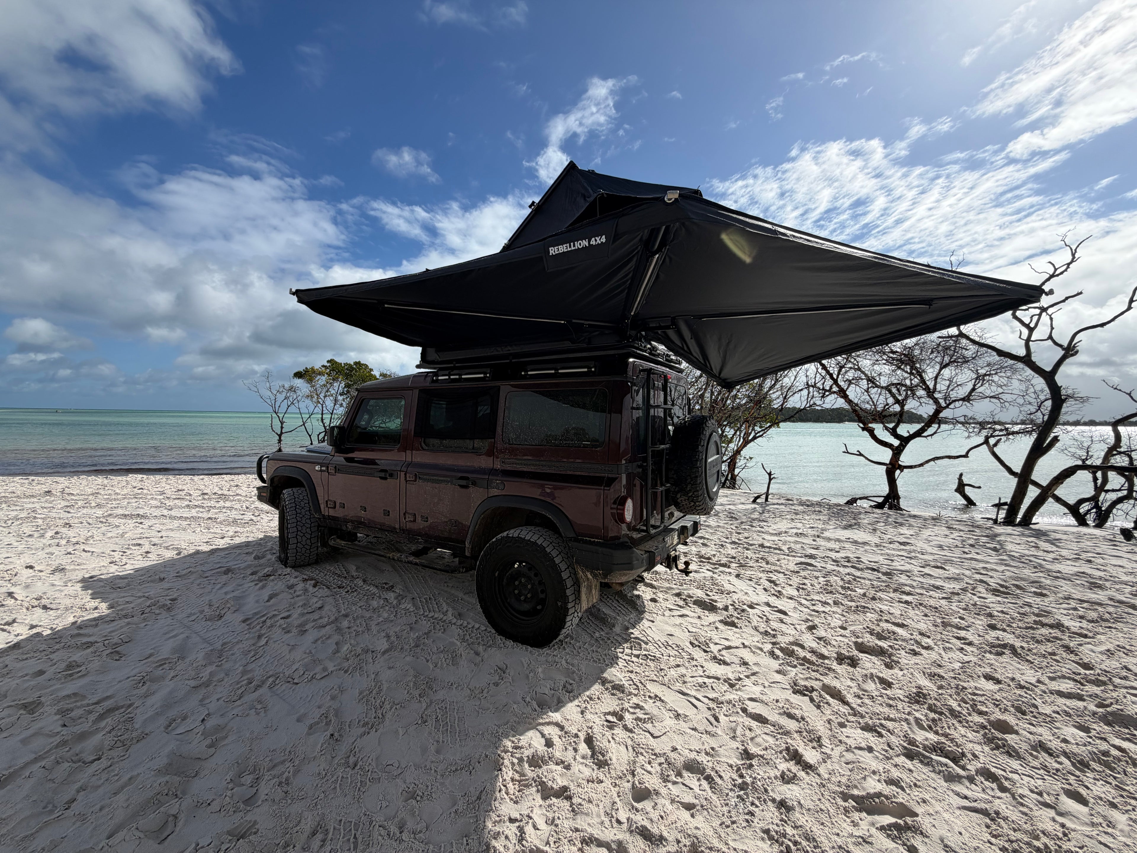 A dark red off-road vehicle parked on a sandy beach with a large black rooftop tent canopy extended over the vehicle, under a blue sky with clouds.
