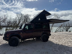 A dark brown Jeep Wrangler parked on sandy ground near a body of water, with a rooftop tent extended above the vehicle under a blue sky with clouds.