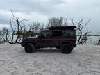 A dark red off-road SUV with a roof top tent is parked on a sandy beach with bare trees and water in the background under an overcast sky.