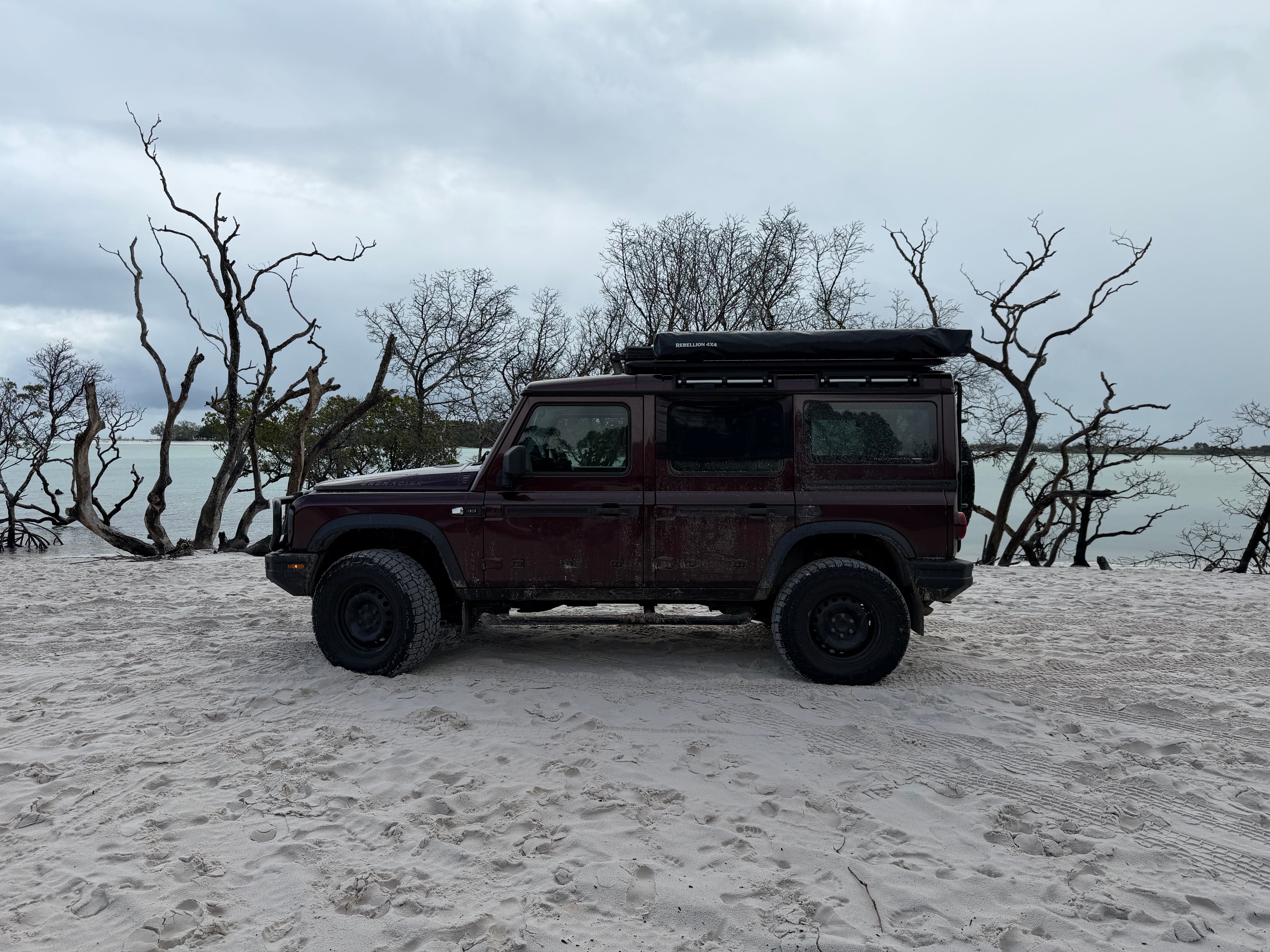 A dark red off-road SUV with a roof top tent is parked on a sandy beach with bare trees and water in the background under an overcast sky.
