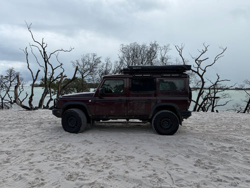 A dark red off-road SUV with a roof top tent is parked on a sandy beach with bare trees and water in the background under an overcast sky.