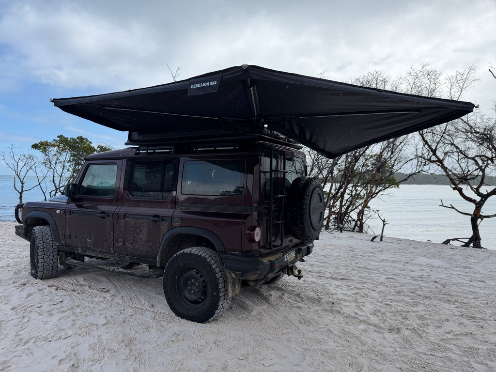 A dark maroon off-road vehicle parked on white sand beach with a large black rooftop awning extended over the vehicle. Bare trees and water are visible in the background under a cloudy sky.