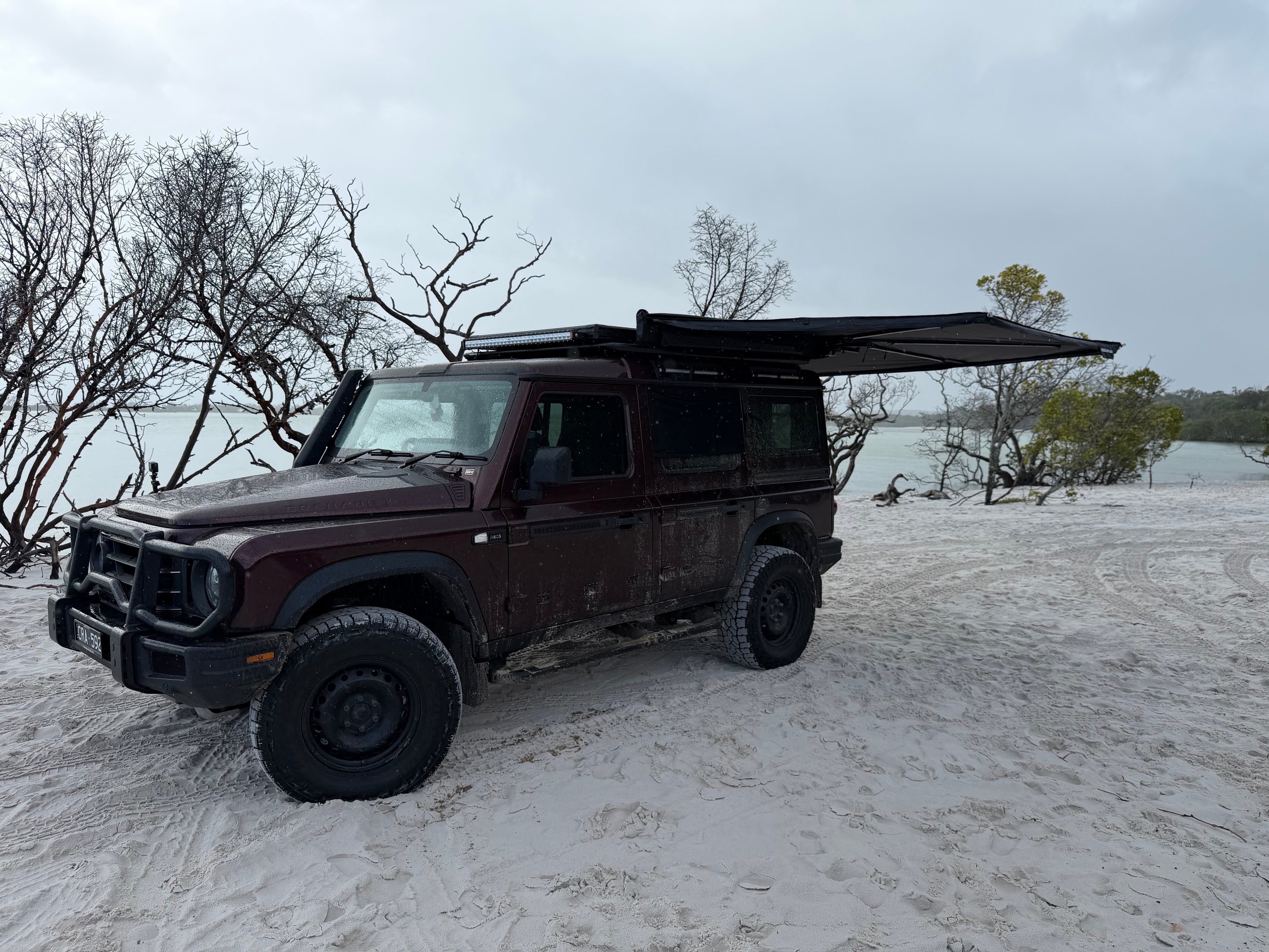 A dark red off-road SUV parked on white sand near water, with a black roof tent awning extended over the rear cabin. Bare trees and an overcast sky are visible in the background.
