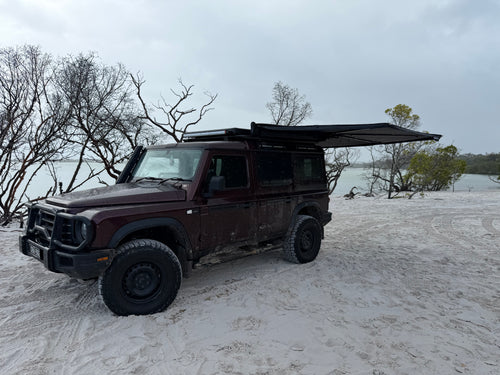 A dark red off-road SUV parked on white sand near water, with a black roof tent awning extended over the rear cabin. Bare trees and an overcast sky are visible in the background.