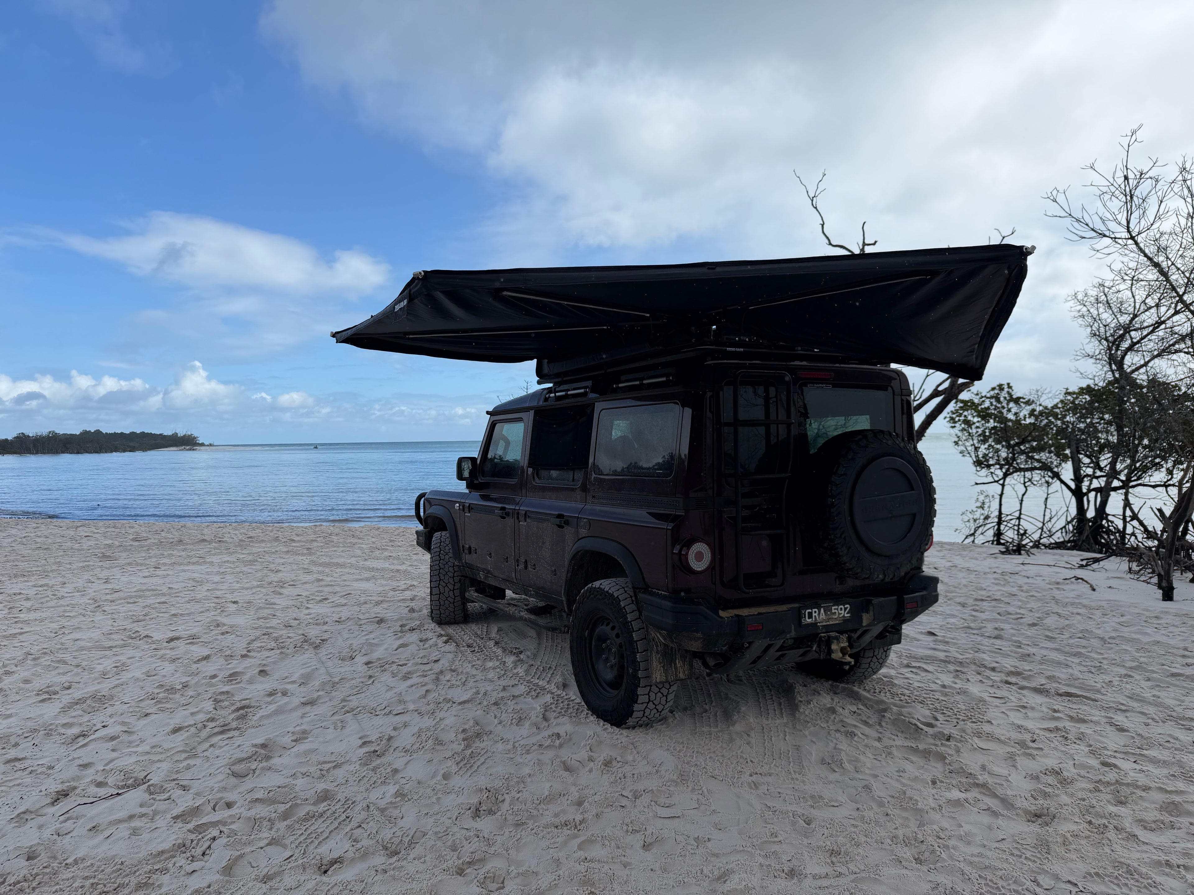 A black off-road vehicle parked on a sandy beach with a large black rooftop canopy extended, overlooking blue water and sky with clouds.