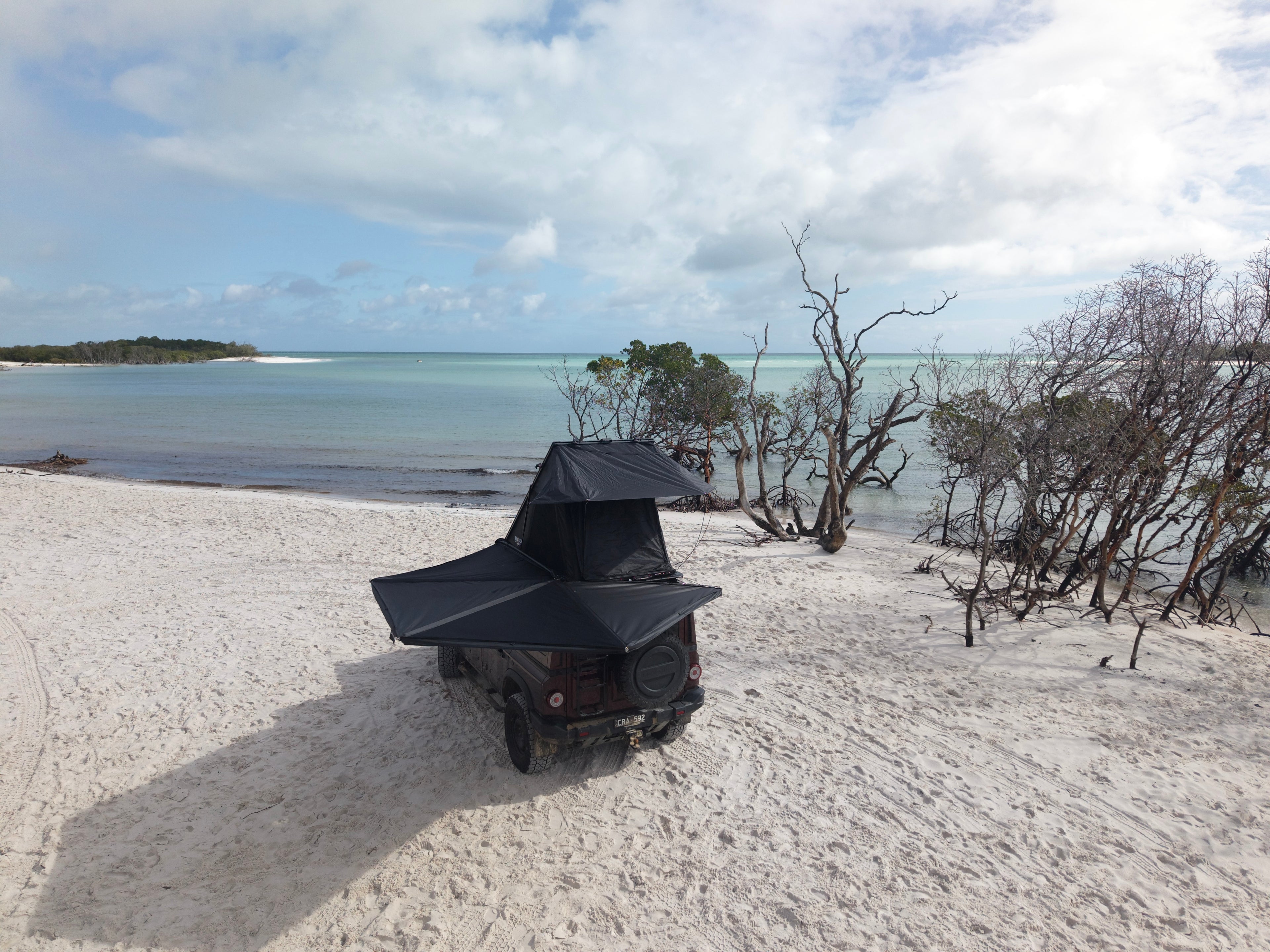 A black rooftop tent mounted on a dark off-road vehicle parked on a sandy beach with ocean, trees, and cloudy sky in the background.