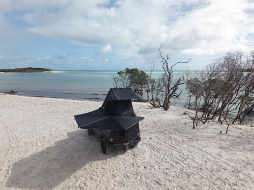 A black rooftop tent mounted on a dark off-road vehicle parked on a sandy beach with ocean, trees, and cloudy sky in the background.