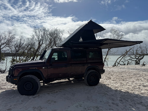 A dark brown Jeep Wrangler parked on sandy ground near a body of water, with a rooftop tent extended above the vehicle under a blue sky with clouds.