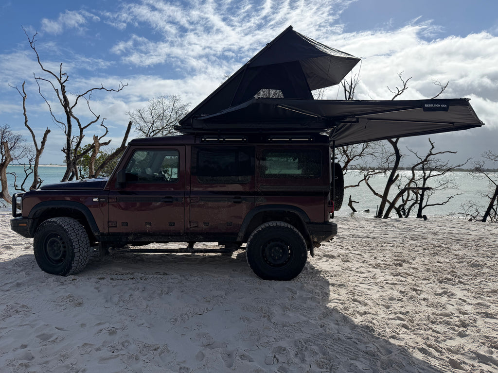 A dark red off-road SUV parked on a sandy beach with a black rooftop tent and extended awning set up above it, with bare trees and water in the background.