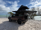 A dark brown off-road SUV parked on a sandy beach with a black rooftop tent and awning deployed above the vehicle, with water, trees, and a blue sky in the background.