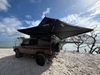 A dark brown off-road SUV parked on white sand beach with a large black rooftop tent canopy extended over the vehicle, under a blue sky with clouds.