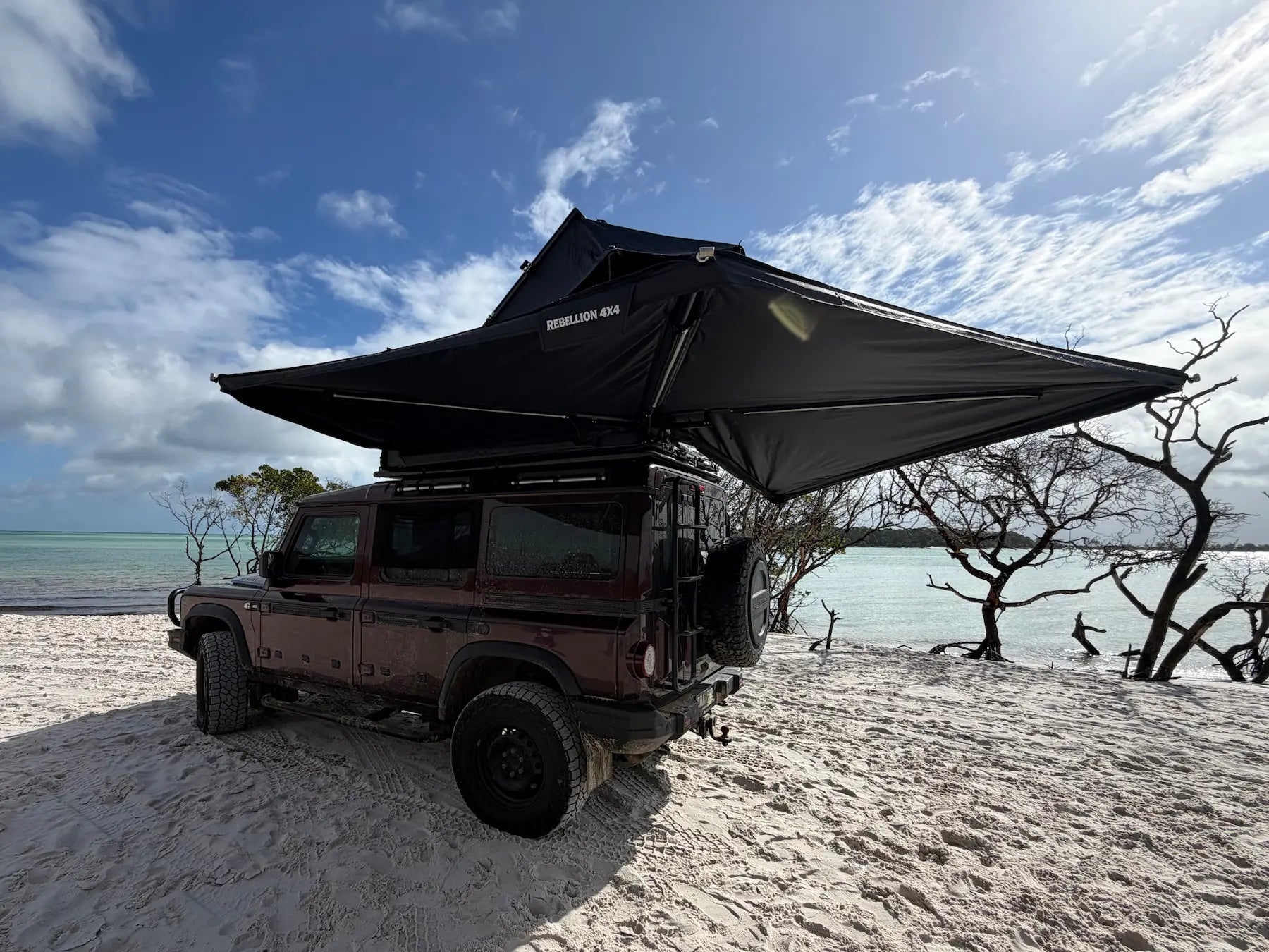 A dark brown off-road SUV parked on white sand beach with a large black rooftop tent canopy extended over the vehicle, under a blue sky with clouds.