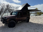 A dark red off-road SUV parked on sandy ground near water, with a rooftop tent and extended awning set up on top. Bare trees and a cloudy blue sky are visible in the background.