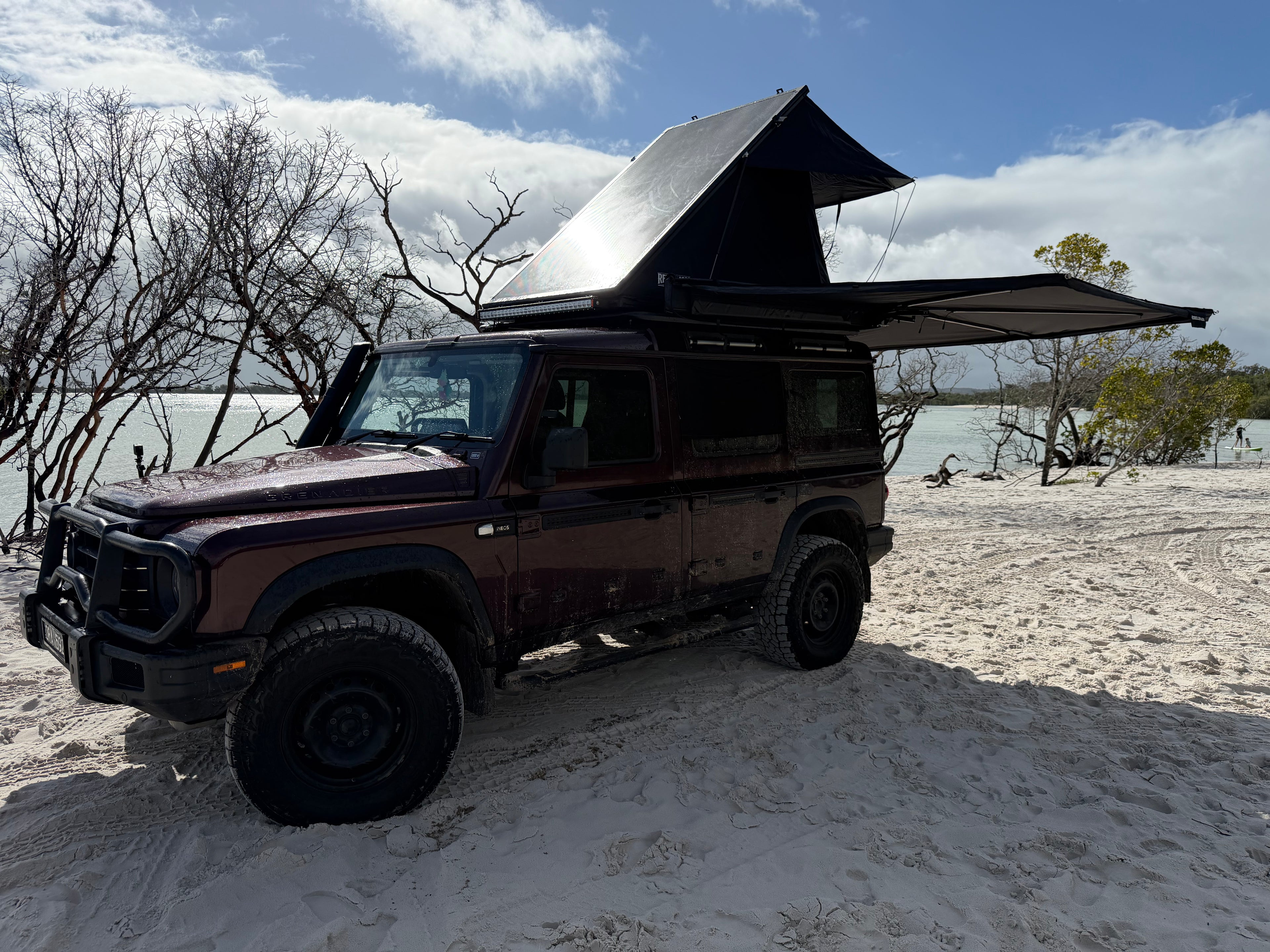 A dark red off-road SUV parked on sandy ground near water, with a rooftop tent and extended awning set up on top. Bare trees and a cloudy blue sky are visible in the background.